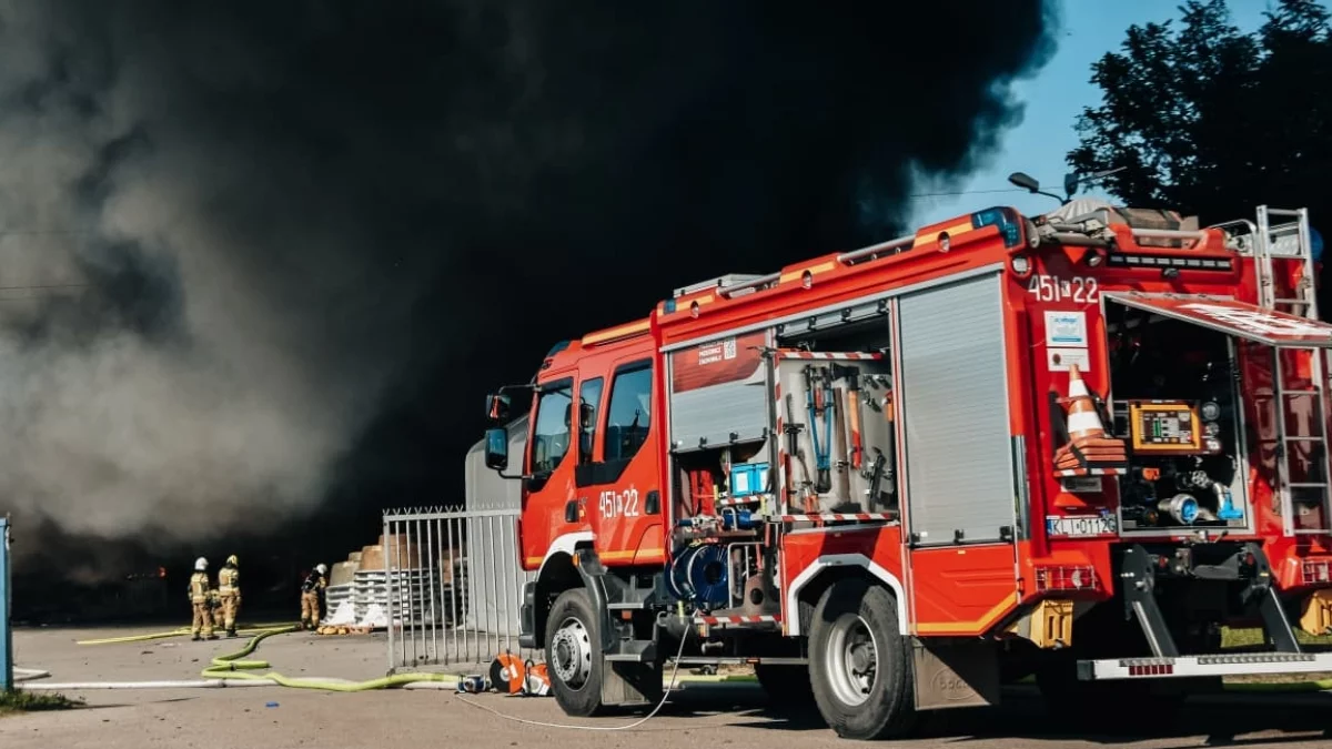 Słup gęstego, czarnego dymu unoszący się nad płonącą halą przemysłową, widoczne wozy strażackie i ratownicy.