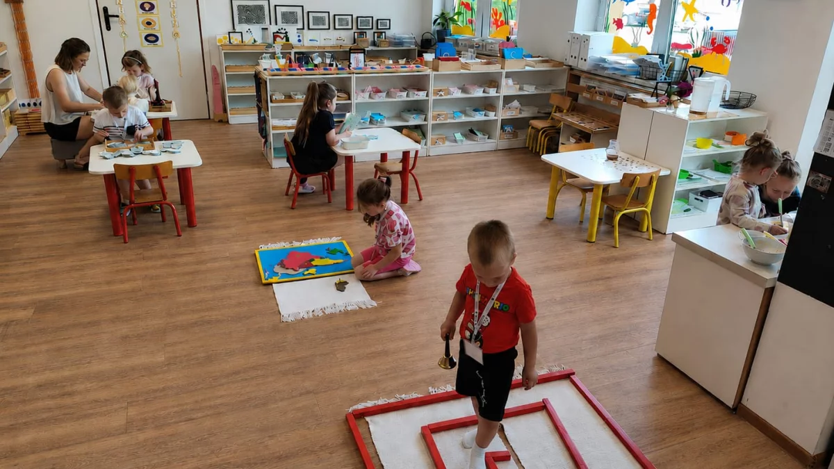 Widok z góry na jasną, przestronną salę przedszkolną Montessori z drewnianą podłogą i otwartymi regałami pełnymi materiałów edukacyjnych. Kilkoro dzieci angażuje się w różne zajęcia. Na pierwszym planie chłopiec w czerwonej koszulce chodzi po labiryncie u